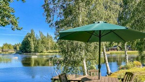 Una terraza de madera con una mesa y sillas, un paraguas verde y vistas a un lago con árboles y un puente al fondo.