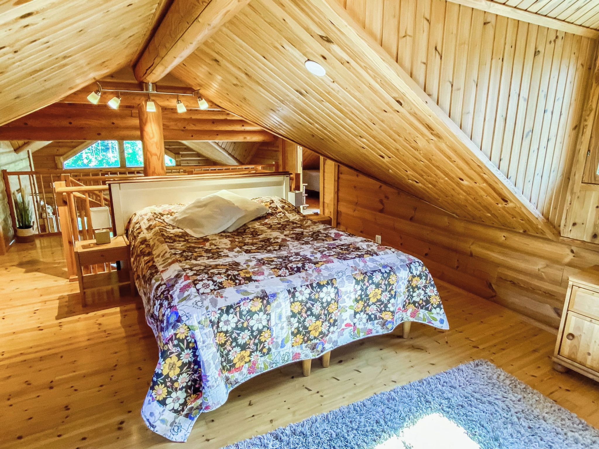 Een slaapkamer met een houten plafond, een bed met een bloemenbedekker en een blauw vloerkleed.