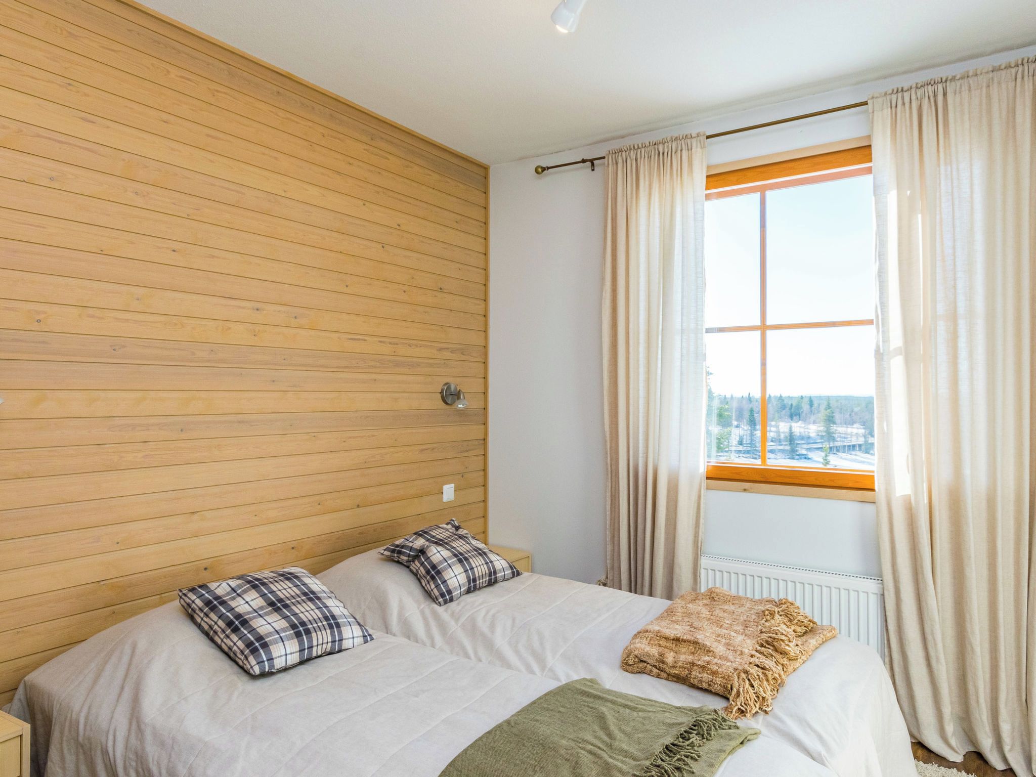 Un dormitorio con cabecera de madera, un ventanal con cortinas, una cama con dos almohadas y una colcha.