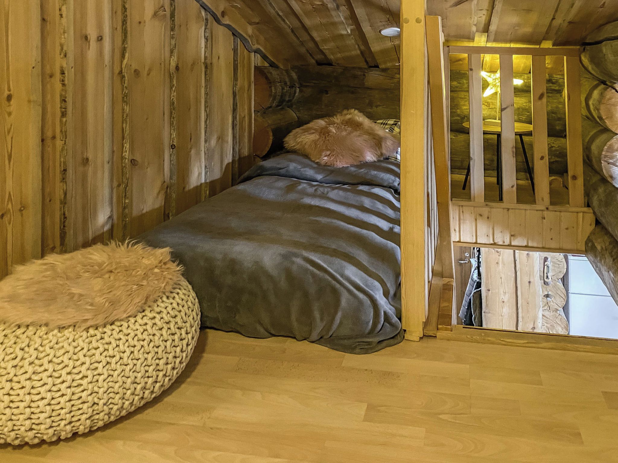A cozy wooden room with a bed, a knitted pouf, and a wooden railing.