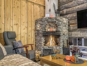 A cozy living room with a stone fireplace, wooden walls, a comfortable sofa, and a flat-screen TV mounted on the wall. - Mäentähti b by Interhome (Kuusamo)