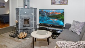 A living room with a stone fireplace, a television displaying a mountain lake, a grey sofa, and a small round table. - Alppitalo punatähti 7 as 2 by Interhome (Nilsiä)
