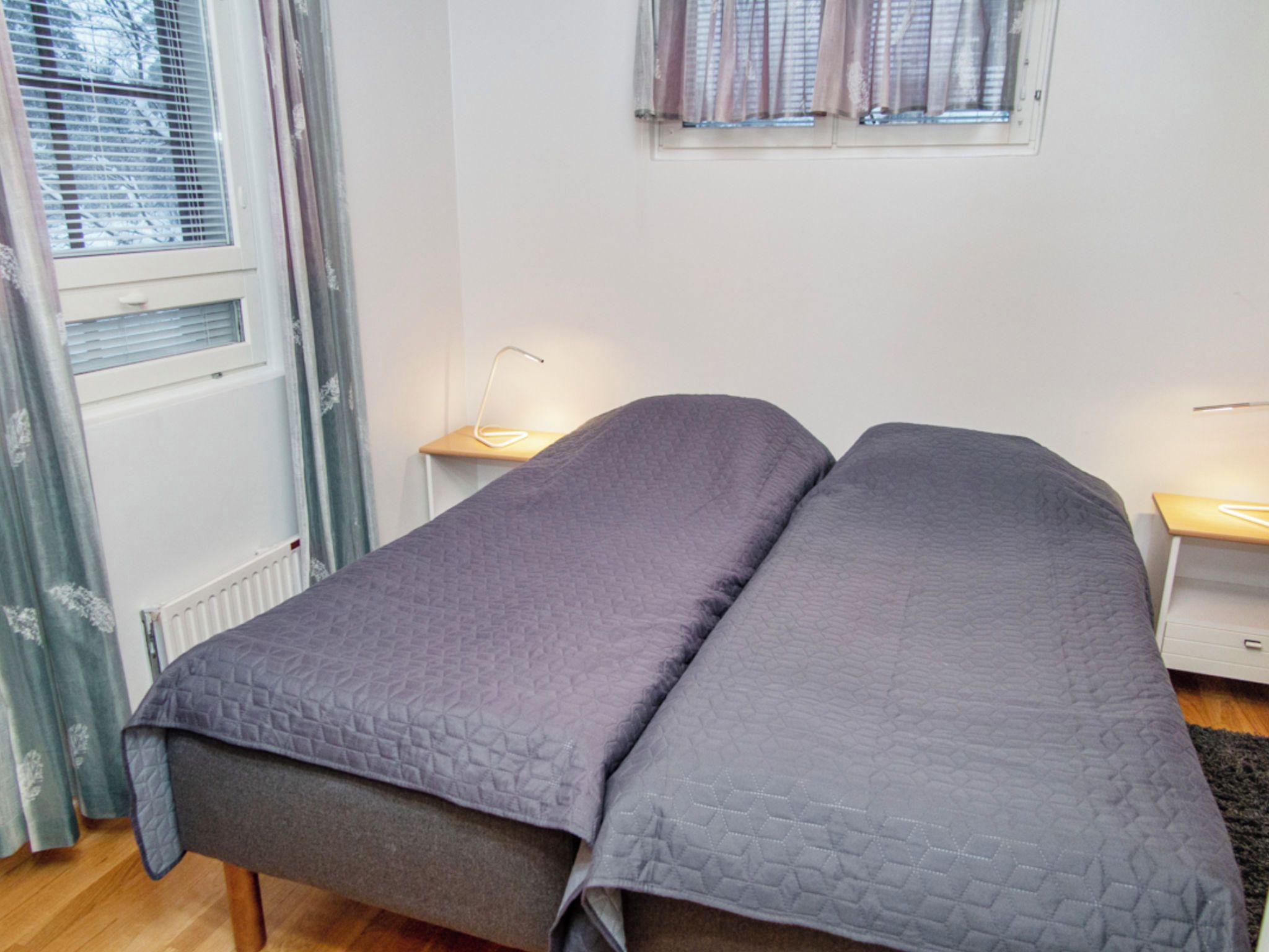 A single bed with a quilted cover in a room with a radiator, a window with curtains, and a bedside table with a lamp.