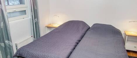 A single bed with a quilted cover in a room with a radiator, a window with curtains, and a bedside table with a lamp.