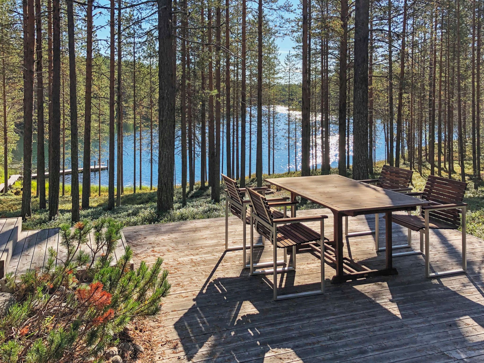 A wooden deck with a table and chairs overlooking a lake surrounded by tall trees.