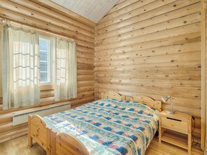 A wooden cabin bedroom with a bed, a nightstand, a lamp, and a window with curtains.