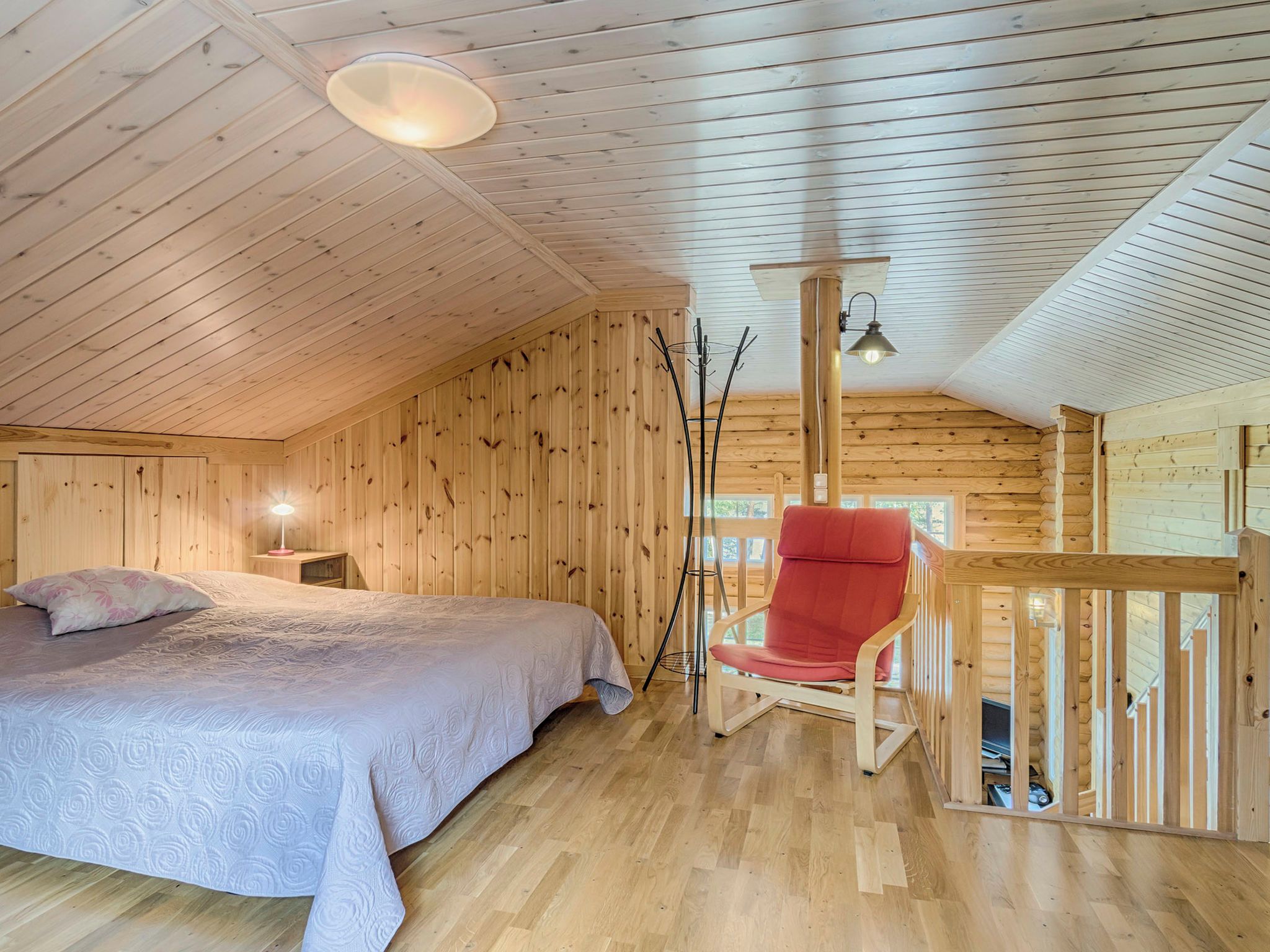 A cozy wooden cabin room with a bed, a red chair, and a small table with a lamp.