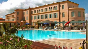 Una amplia piscina con sillones de playa y sombrillas, rodeada por un edificio con múltiples ventanas y balcones.