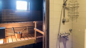 A sauna with wooden benches and a metal bucket, a window with natural light, and a shower area with a metal rack.