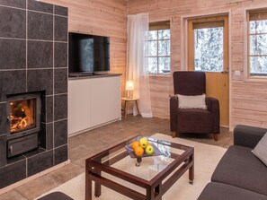A cozy living room with a fireplace, a flat-screen TV, a comfortable armchair, and a coffee table with fruit.