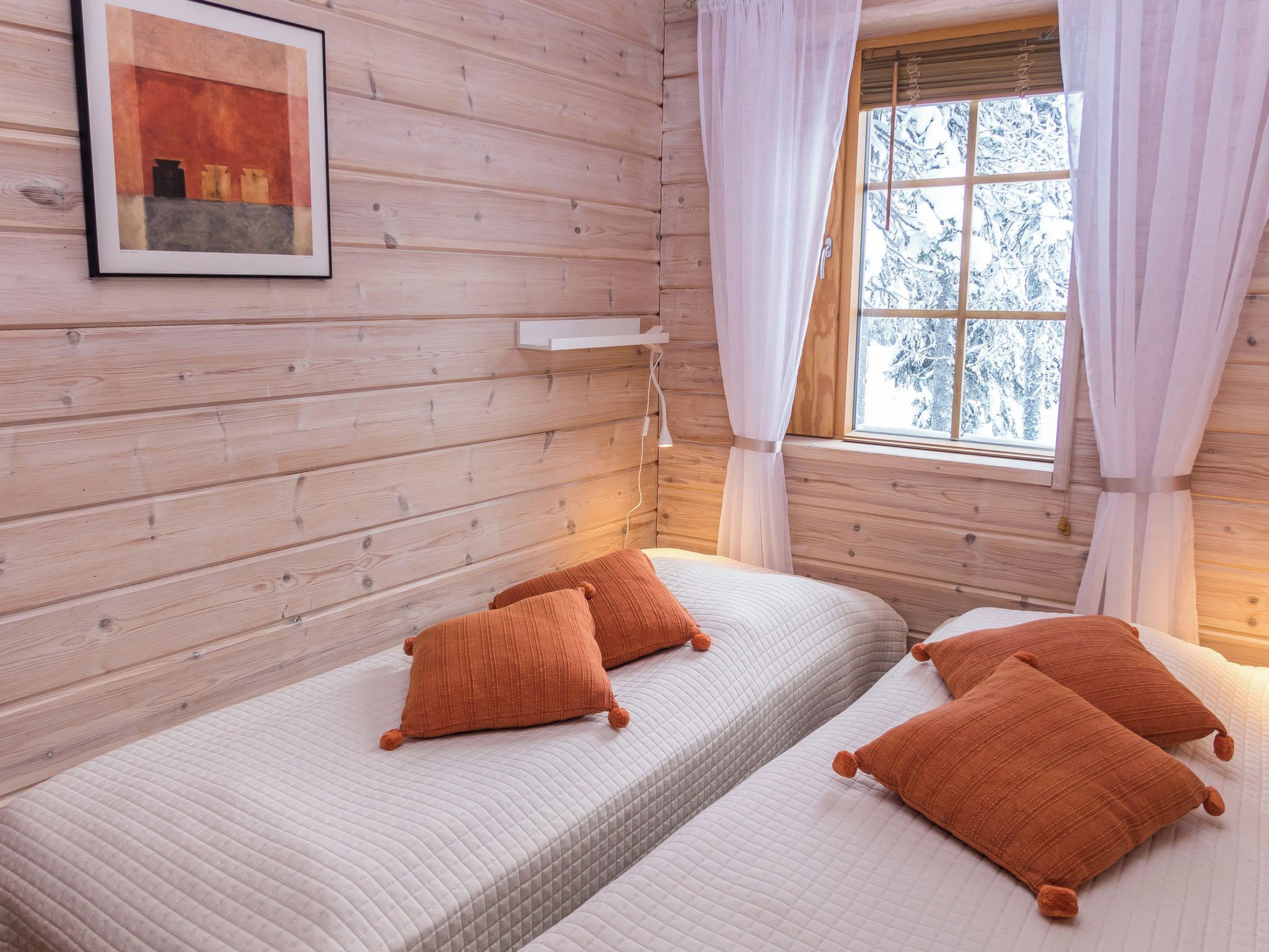 A cabin-style room with wooden walls, a bed with white bedding and orange pillows, a bedside table, and a window with curtains.