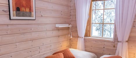 A cabin-style room with wooden walls, a bed with white bedding and orange pillows, a bedside table, and a window with curtains.