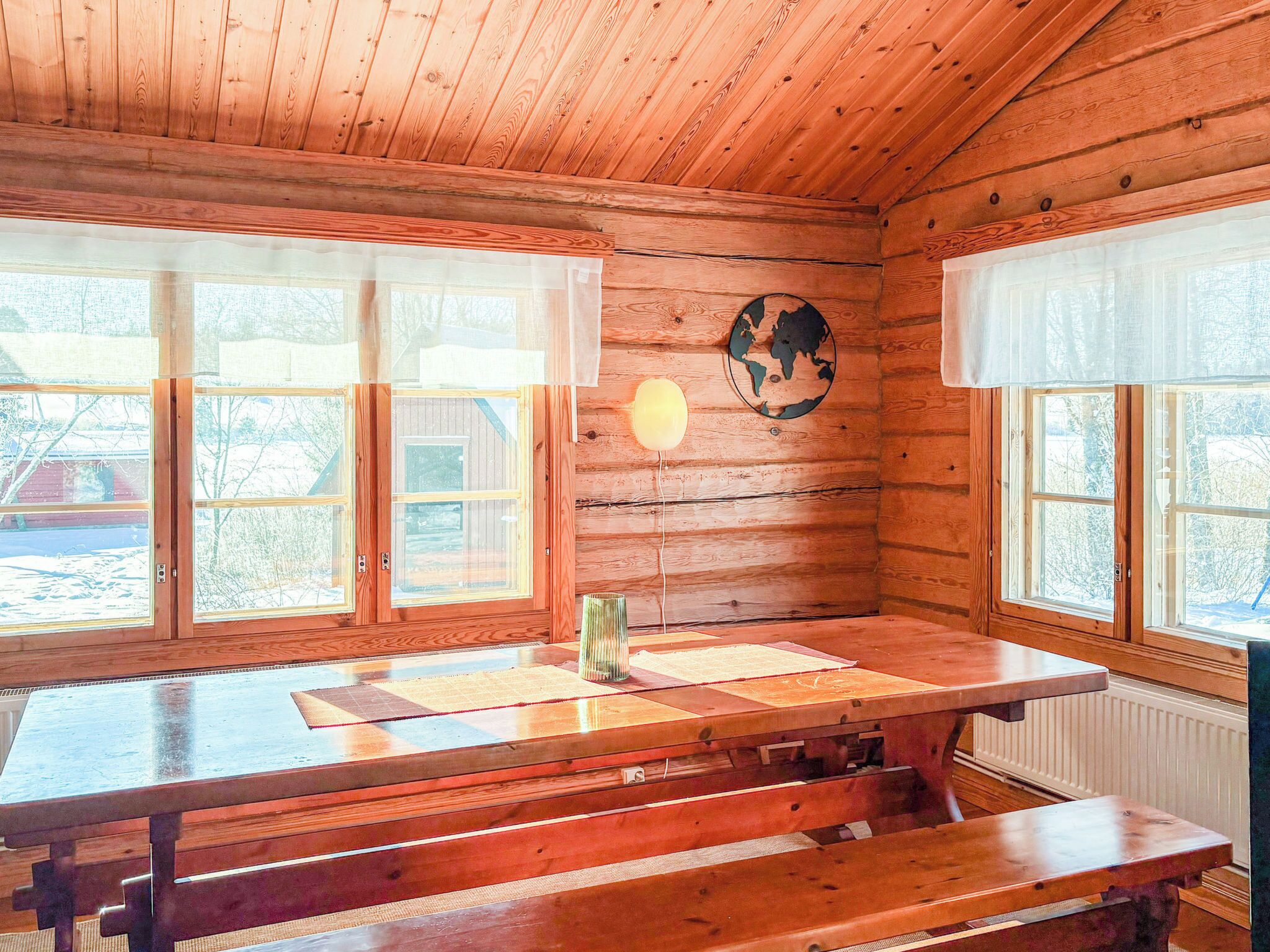 Interior de una cabaña de madera con una gran mesa de madera y bancos, ventanas amplias y un globo decorativo en la pared.