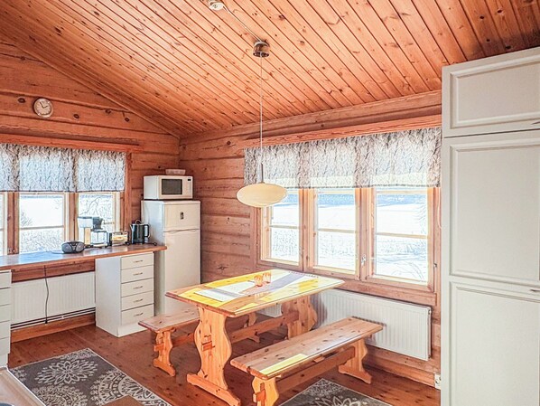 Interior de cabana com paredes de madeira, uma mesa de madeira, bancos, micro-ondas, geladeira e janelas amplas.
