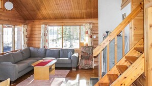 A cozy cabin interior with a wooden staircase, a gray sectional sofa, a small wooden table, and a view of the forest through large windows. - Mäntyniemi by Interhome (Konnevesi)