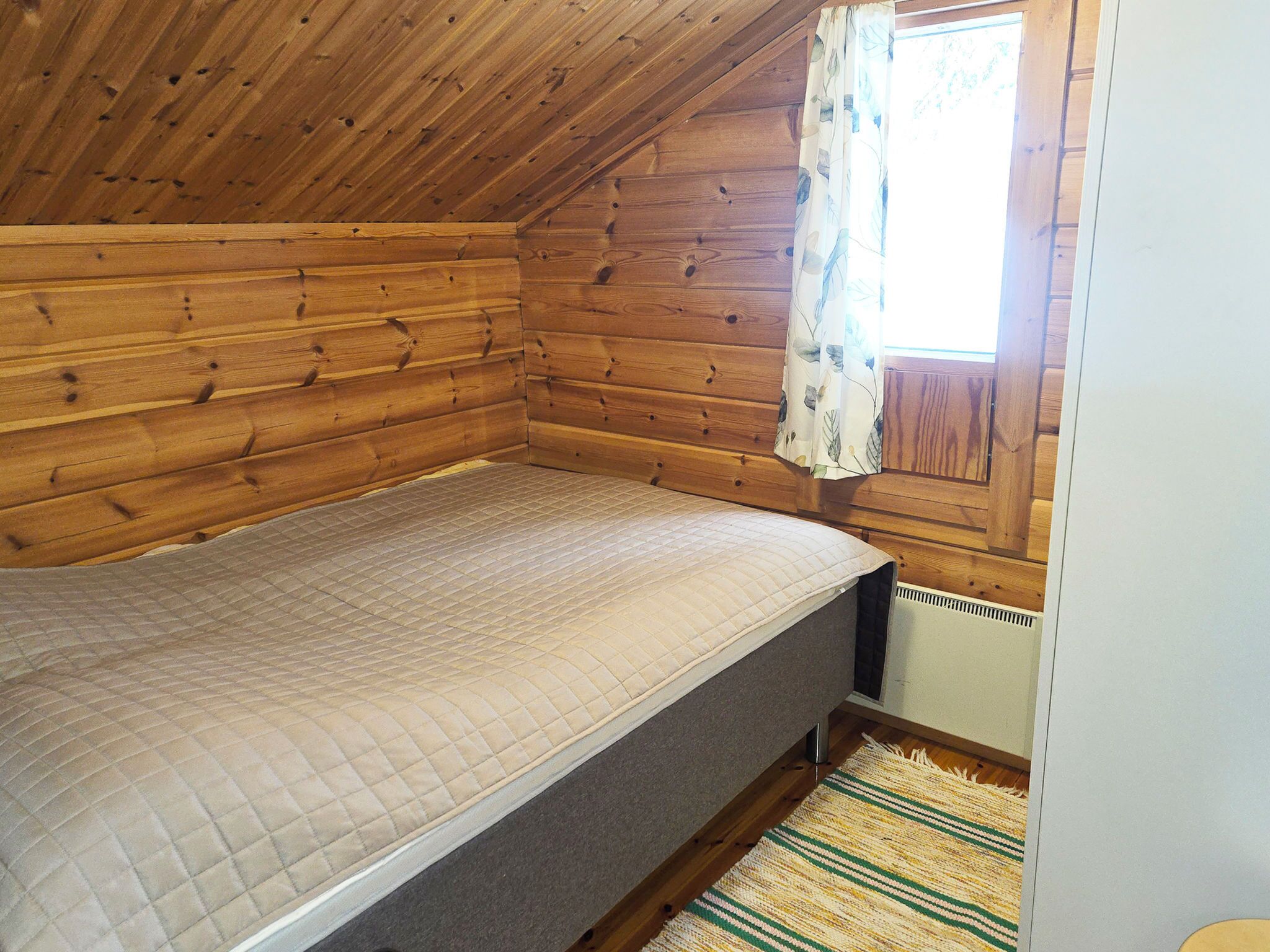 A small, wooden cabin room with a single bed, a window with a curtain, and a radiator.