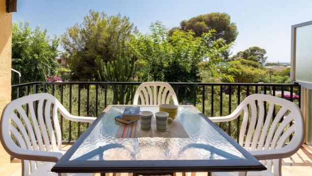 Mesa com tampo de vidro e quatro cadeiras brancas numa varanda com guarda-corpo de metal, com vista para um jardim com diversas plantas e árvores.