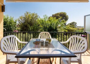 Une table à plateau en verre, entourée de quatre chaises blanches, se trouve sur un balcon doté d’une rambarde en métal, et offre une vue sur un jardin où poussent diverses plantes et arbres.