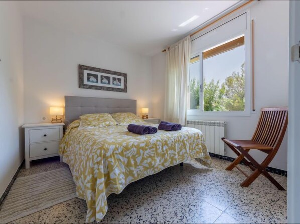 Une chambre à coucher avec un lit, deux tables de chevet, une fenêtre avec des rideaux, une chaise en bois et un tableau au mur.