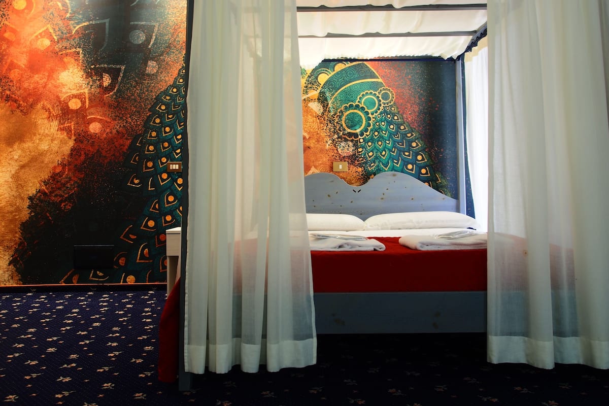 Una habitación con una cama, cortinas blancas y un tapiz colorido en la pared.