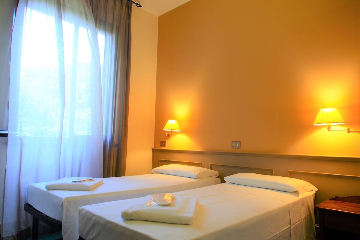 Habitación de hotel con dos camas, cada una con una almohada y una toalla doblada. Dos lámparas de pared proporcionan luz.