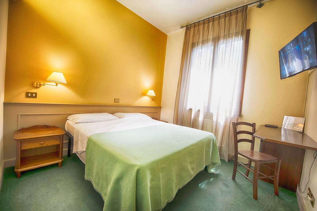 Habitación de hotel con cama, mesita de noche, lámpara, silla, escritorio y televisión en la pared.