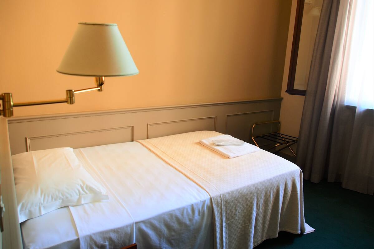 Una habitación de hotel con una cama, una lámpara, una silla y un ventanal con cortinas.