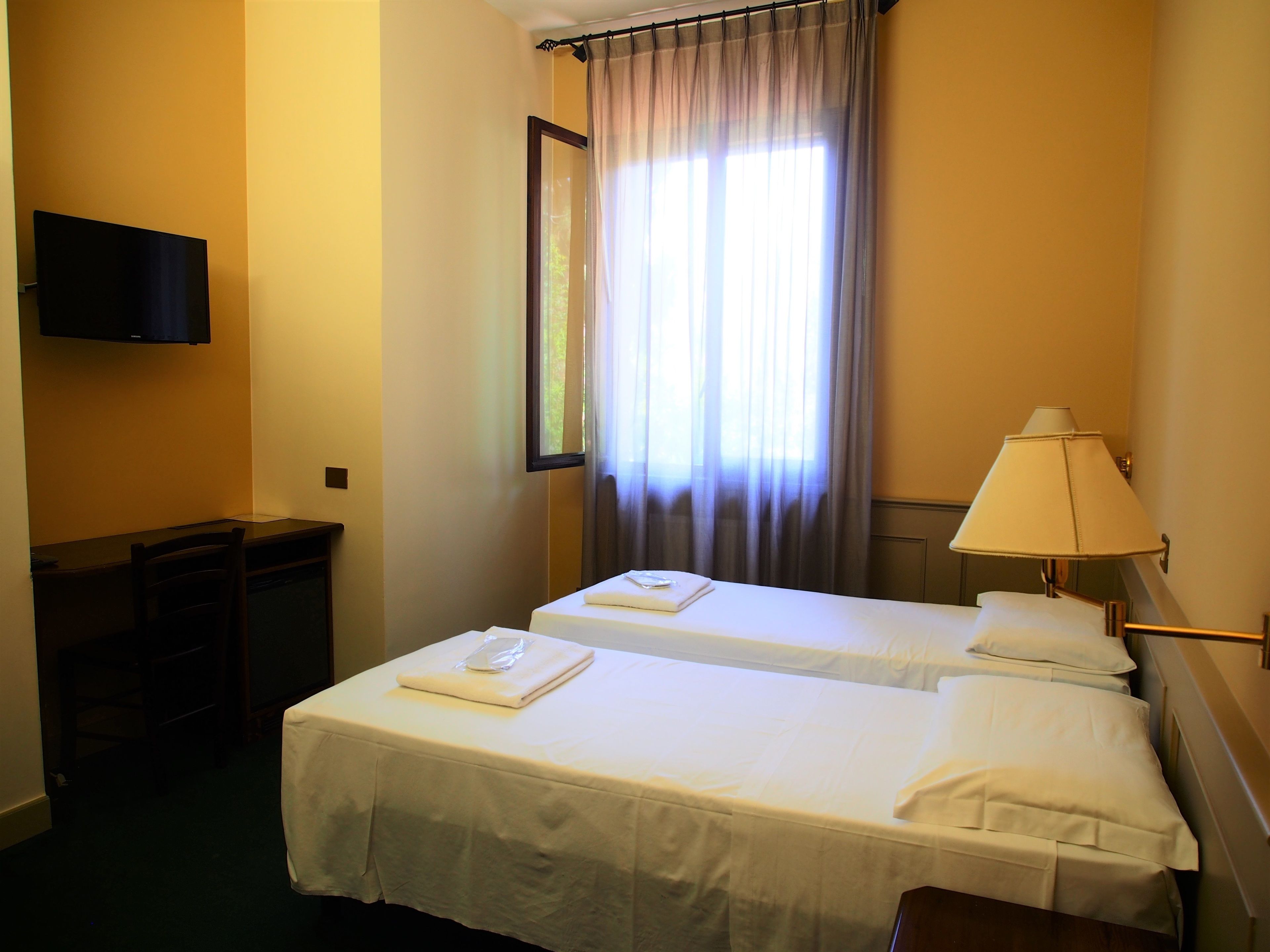 A hotel room with two single beds, a desk, a chair, a TV mounted on the wall, and a window with curtains.