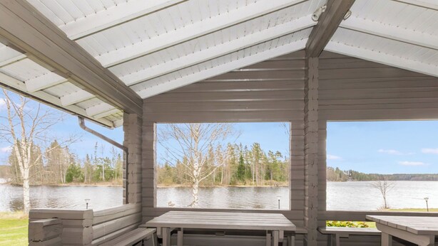 Un área de picnic techada con bancos y una mesa, con vista a un lago rodeado de árboles y una orilla distante.