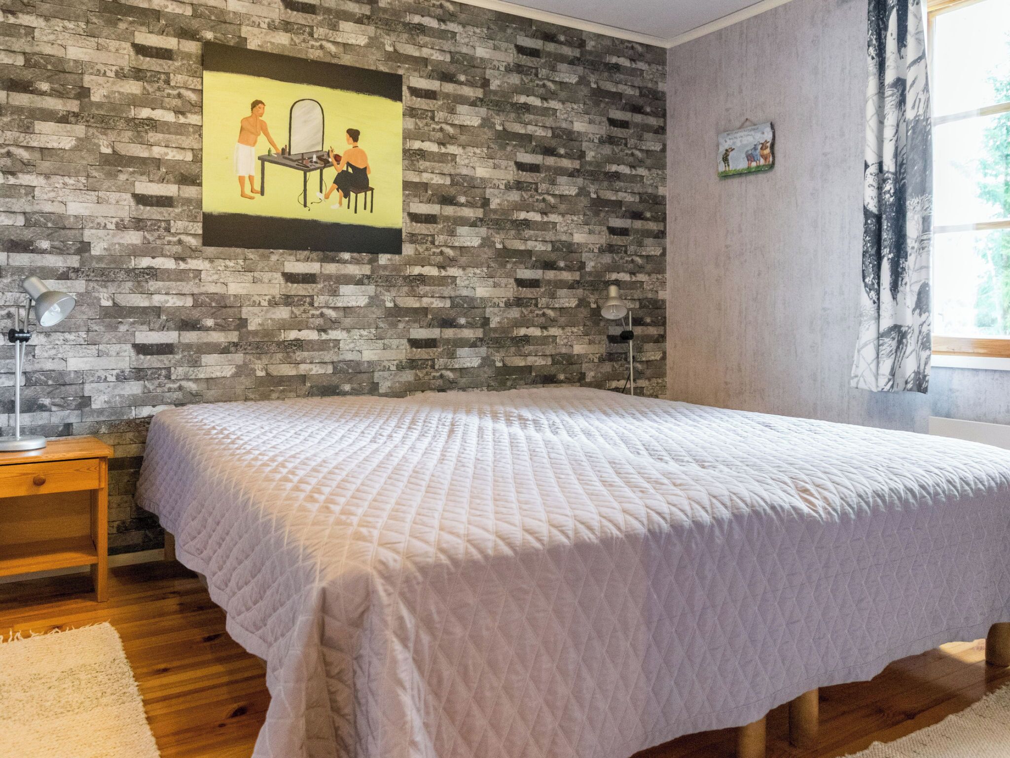 A bedroom with a bed, a nightstand, a lamp, a brick-patterned wall, and a window with curtains.