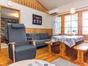 Une pièce avec un plafond en bois, un canapé gris, une chaise en bois, une petite table recouverte d’une nappe à motifs et une horloge au mur.