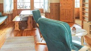 Una habitación acogedora con paredes y suelos de madera, una butaca tapizada de verde, una mesa de madera y una ventana con cortinas.