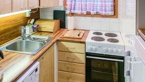 A compact kitchen with wooden cabinets, a sink, a stove, and a window with a plaid curtain. - Pikku-metso by Interhome (Kuusamo)