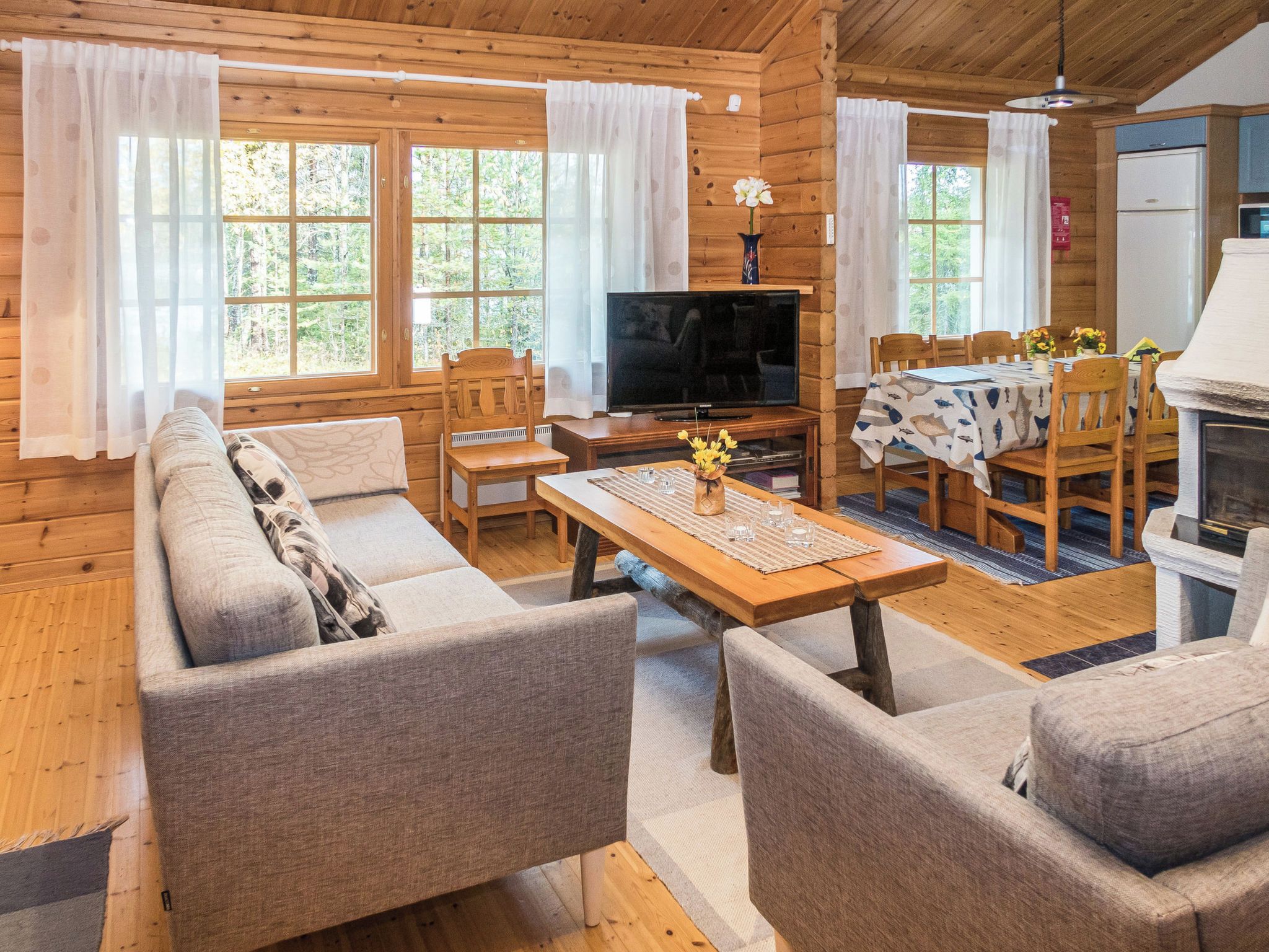 Ein gemütliches Blockhauss-Interieur mit einer Couch, Sesseln, einem Esstisch, einem Fernseher und einem Kamin.