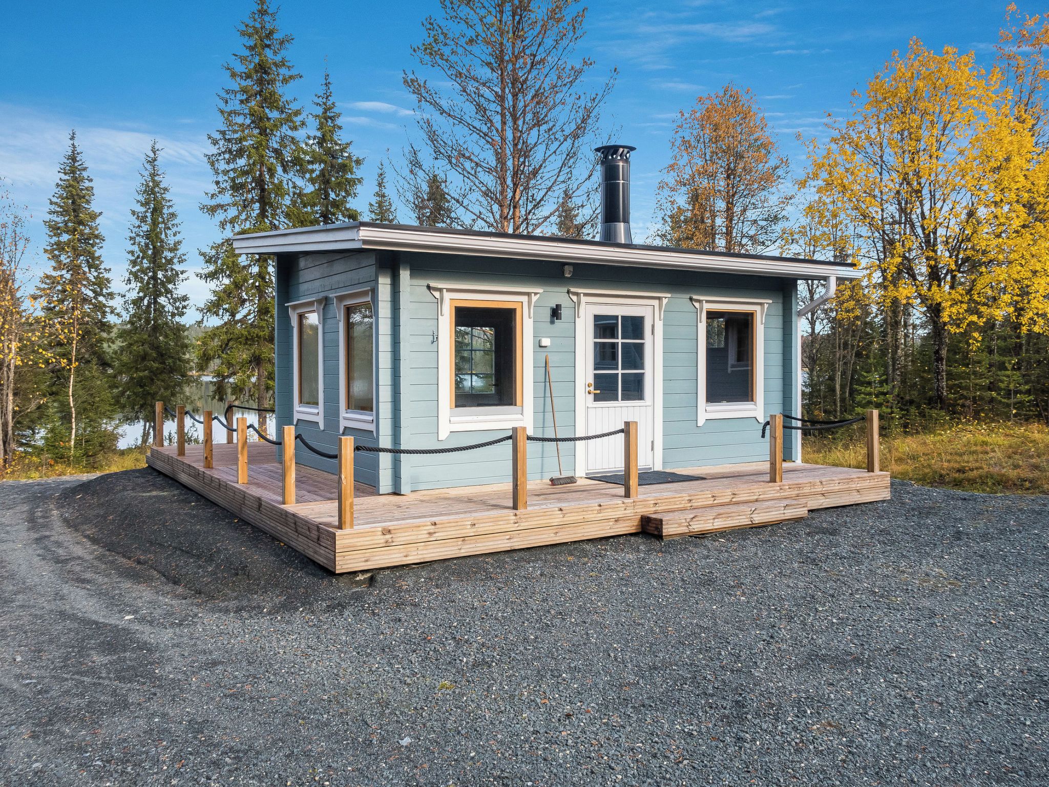 Eine kleine, eingeschossige Holzhütte mit einer Holzterrasse, umgeben von Bäumen und einem Kiesweg.