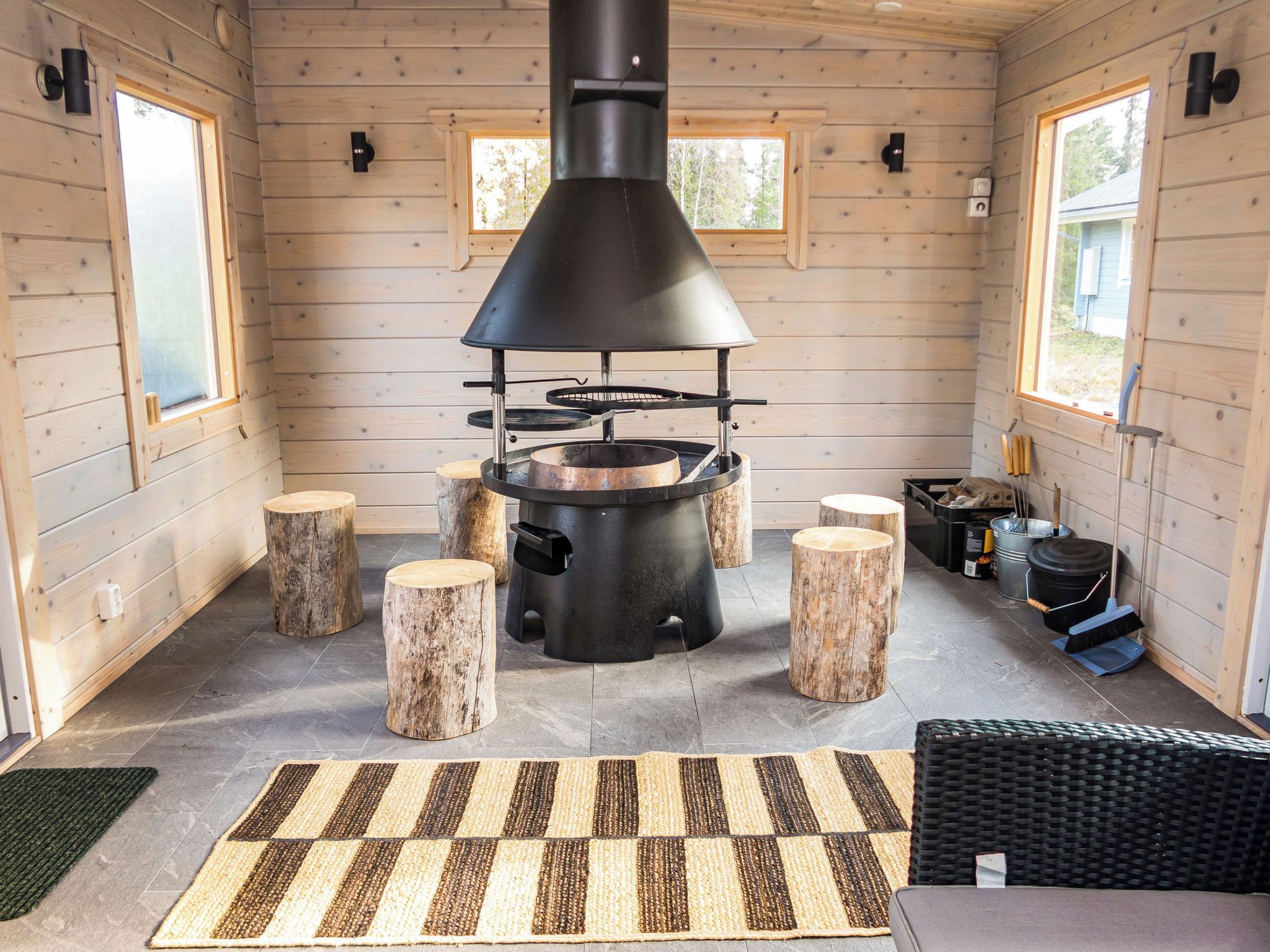 A rustic cabin interior with a wood-burning stove, wooden stools, and a woven rug.