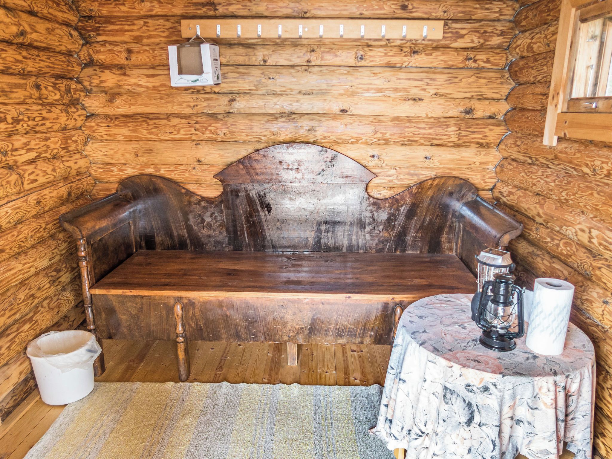 A rustic wooden cabin interior with a bench, a small table with a lantern, and a hanging light fixture.