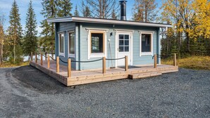 A small, single-story cabin with a chimney, surrounded by a wooden deck and a gravel path.