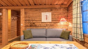 Ein gemütliches Blockhaus-Interieur mit Holzbalkendecke, einem grauen Sofa mit grünen Kissen, einem Korbwäschesammler und einem gerahmten Bild an der Wand.
