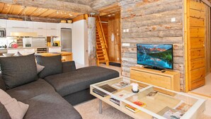 A log cabin interior with a modern kitchen, a living area with a sofa and coffee table, and a TV displaying a mountain landscape.