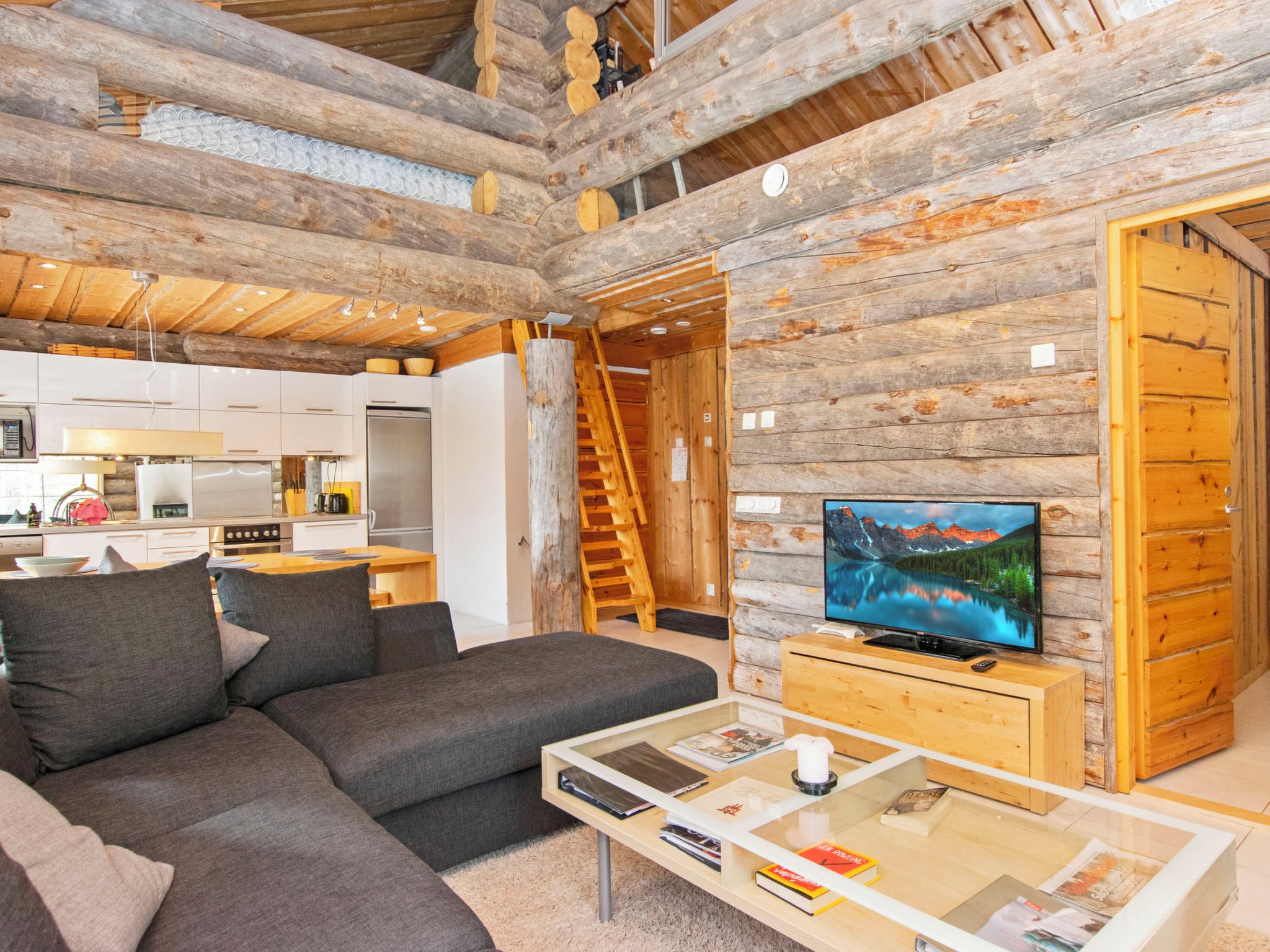 A modern log cabin interior with a living area, kitchen, and a large TV screen displaying a scenic landscape.