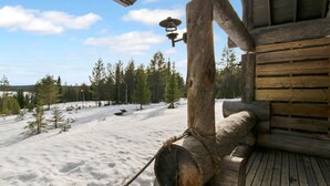 A log cabin with a wooden deck and a rope railing overlooking a snowy landscape with trees.