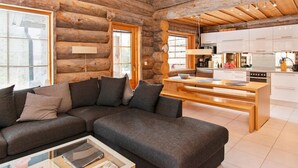 A log cabin interior with a living area featuring a sofa and a coffee table, a kitchen with modern appliances, and a wooden ceiling.