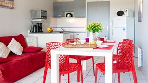 Un espace de vie compact comprenant un coin repas avec une table blanche et des chaises rouges, un canapé rouge et une cuisine équipée d’appareils électroménagers modernes.