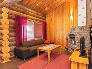 Interior de una cabaña de troncos con chimenea de piedra, un sofá, una mesa y una ventana con cortinas.