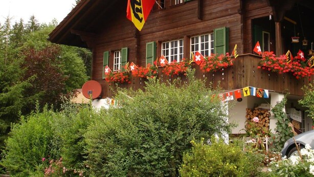 Un chalet en bois doté d’un toit à pente, de nombreuses fenêtres avec des volets verts, et d’un balcon agrémenté de fleurs rouges et de drapeaux suisses.