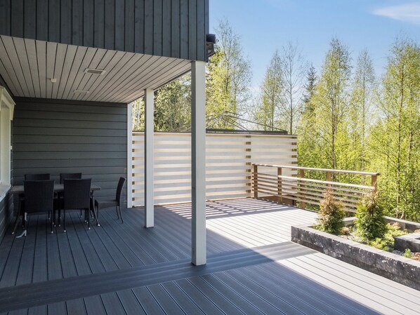 A covered outdoor deck with a dining area, a railing, and a view of trees.