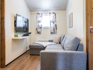 Un espace de vie compact comprenant un canapé gris, un téléviseur à écran plat fixé au mur, une petite étagère avec des appareils électroniques et une fenêtre agrémentée de rideaux à motifs.