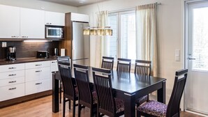 A modern kitchen with white cabinets, a microwave, and a refrigerator. There is a dining table with six chairs and a chandelier.