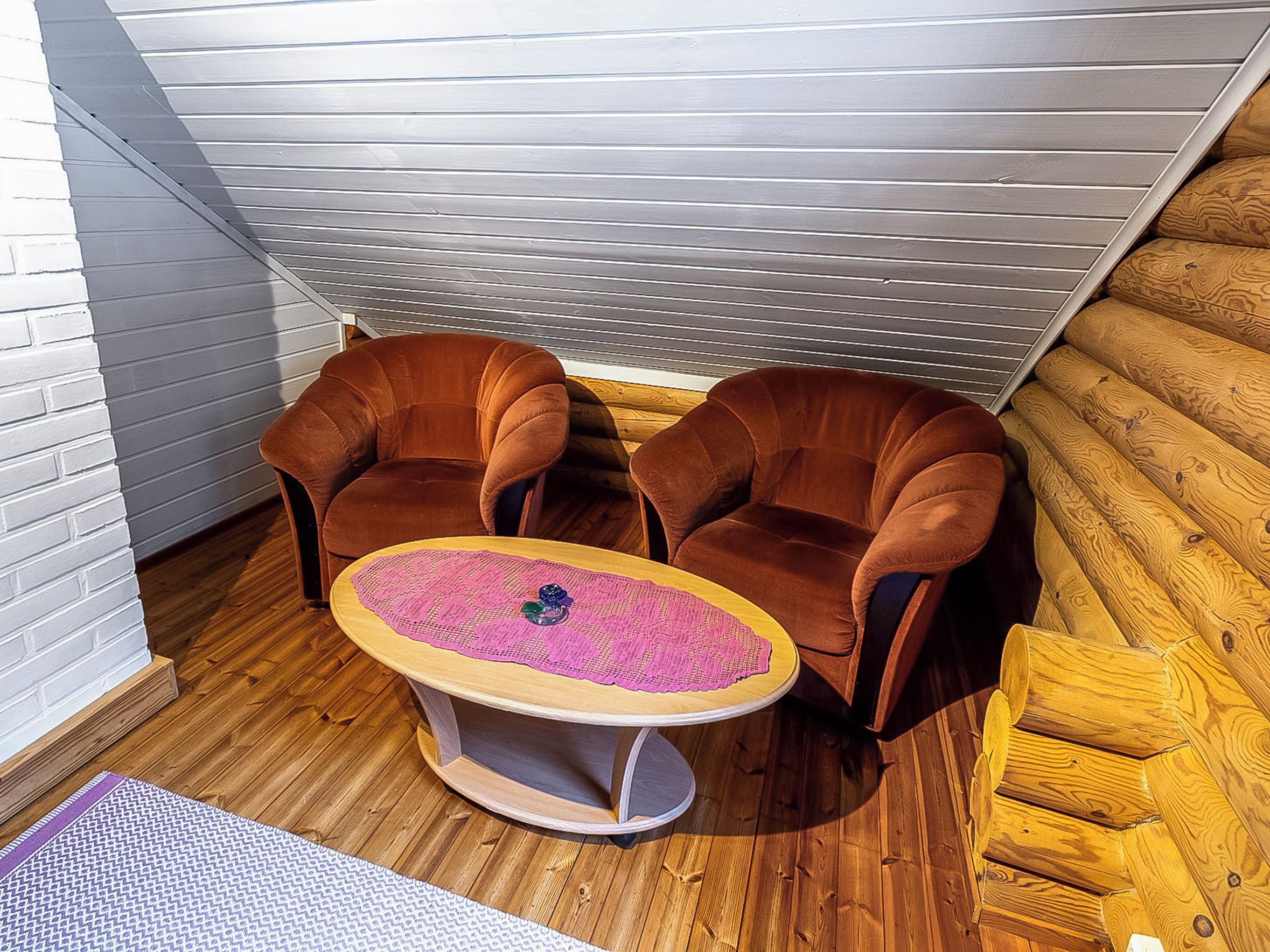 Een hoekzaak met twee bruine fauteuils, een klein rond tafeltje met een roze tafelkleed en houten vloeren.